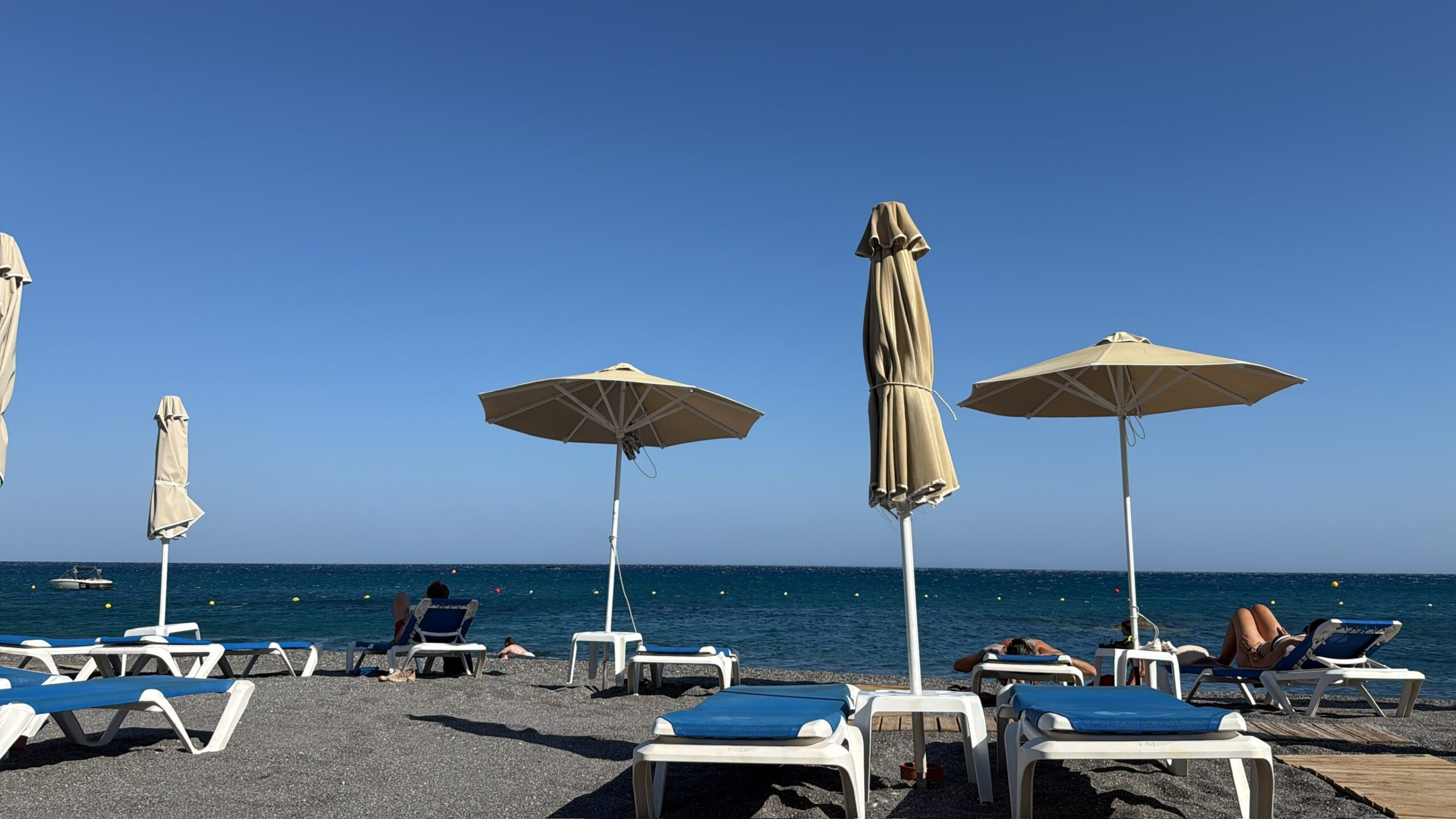 Sonnenliegen und helle Sonnenschirme am Kiesstrand des ROBINSON Ierapetra vor dem blauen Meer.